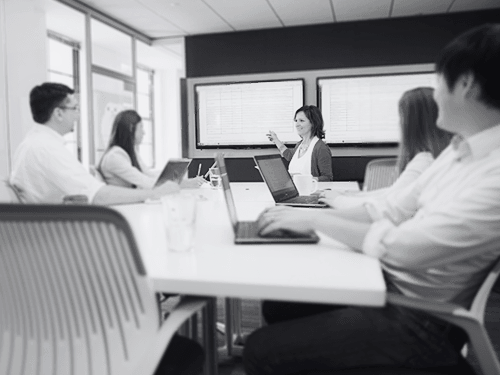 Five people at a board table; one of the women is presenting using the screen behind her
